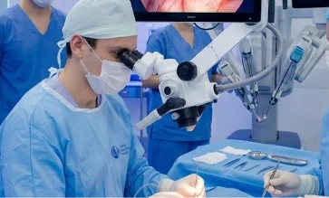 Urologista usando microscópio cirúrgico durante microcirurgia, com monitor ao fundo